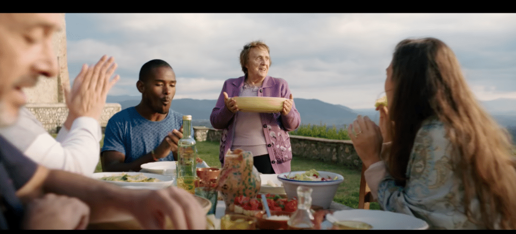 Screenshot of an idyllic scene: people sit at the table, Italian nonna smilingly brings the food.